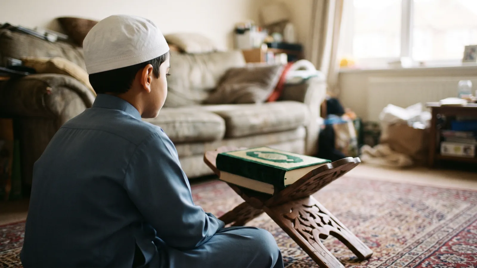Boy in a white kufi sitting on a rug at home with a Quran on a wooden stand